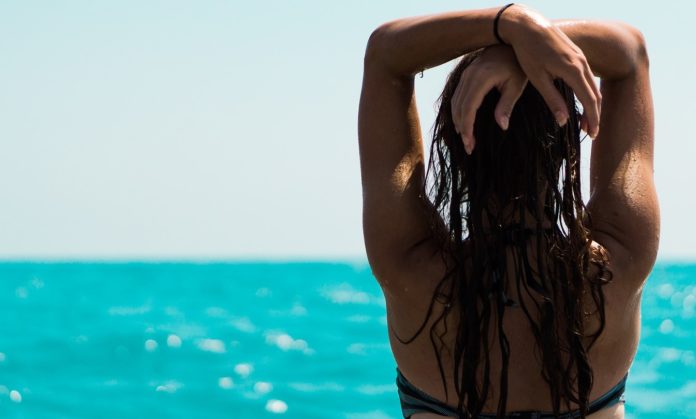 Estate e Turismo - Una donna di schiena in bikini con le braccia alzate e le mani sopra la testa, con lunghi capelli neri guarda il mare blu