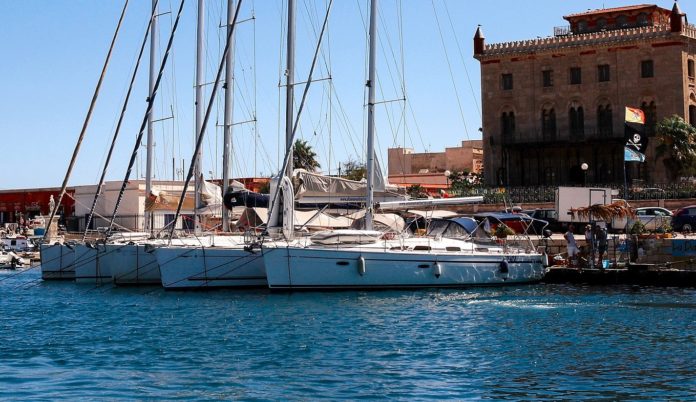 Festivalflrio all'isola di Favignana, nella foto il porto dell'isola con tante barche ormeggiate sul mare blu, e sullo sfondo le coste e una grande casa a più piani