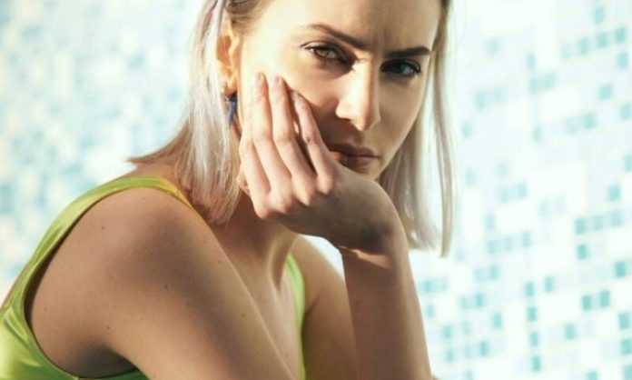 Laura Di Lenola in foto ha uno sfondo di piscina dietro di sé, in primo piano, guarda la fotocamera, ha una mano appoggiata alla guancia, è bionda