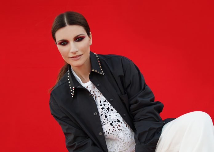 Laura Pausini, con uno sfondo rosso, è inginocchiata con una gamba solvata, indossa una maglia e un pantalone bianchi, uno stivale nero, una giacca nera, ha la coda e un trucco fucsia sugli occhi