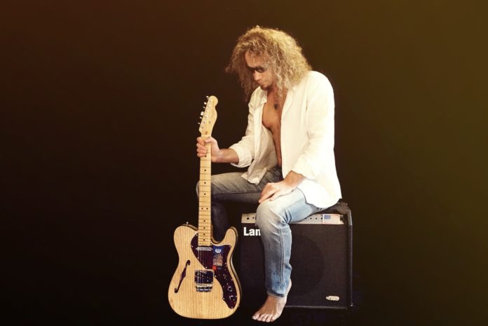 maurizio ferrandini seduto su uno sgabello, con una chitarra elettrica appoggiata al pavimento. capelli biondi, indossa jeans chiari e una giacca bianca