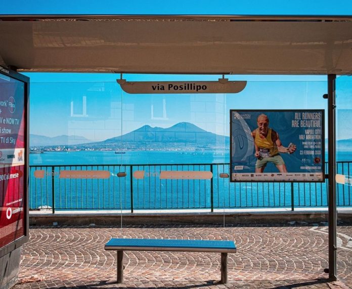 Via Posillipo, fermata bus. Fermata dell'autobus con la pensilina trasparente. Dietro è possibile vedere il Vesuvio e il mare . Sopra la panchina una targhetta con il nome della fermata Via Posillipo.