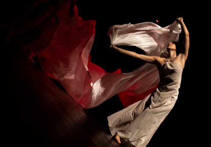 Danza Tra Dante e MIoto Greco - una ballerina vestita di bianco fa volteggiare un raso rosso facendo un arco con il suo corpo. E' bellissima e danza nel buio illuminata solo da uno spot bianco