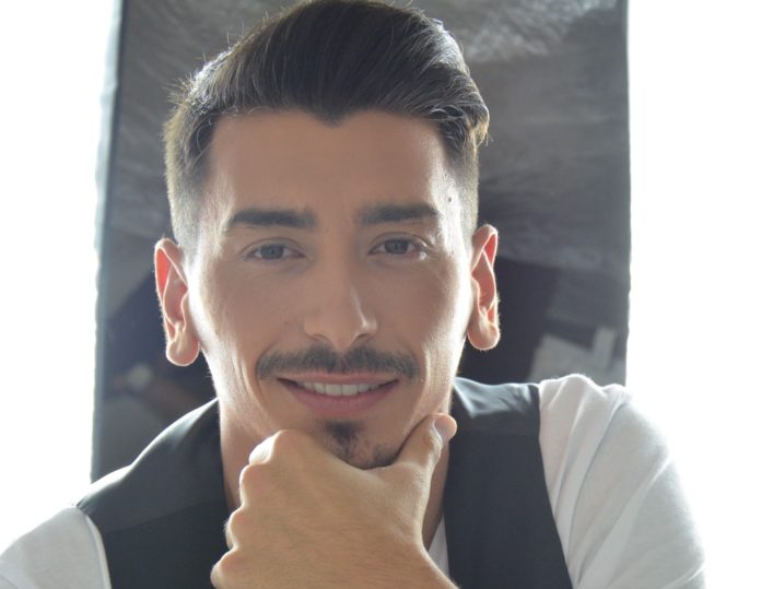 AStefano de Filippo in un primo piano sorridente, concapelli castani corti ai lati e piu lunghi in alto, tirati all'indietro, con baffi e pizzetto, si tiene il mento con la mano sinistra. Indossa una maglia bianca e un gillet nero
