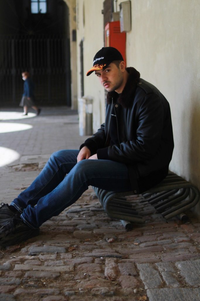 pupille vinile - il giovane cantautore seduto a terra, indossa jeans blu, cappello da baseball e giuppotto di pelle nera con colletto di pelliccia