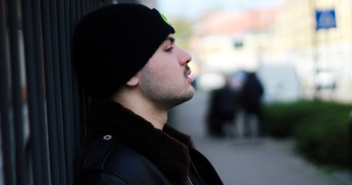 pupille-vinile: il giovane cantautore appoggiato a un muro, indossa giubbotto nero col collo di pelliccia e berretto di lana nero