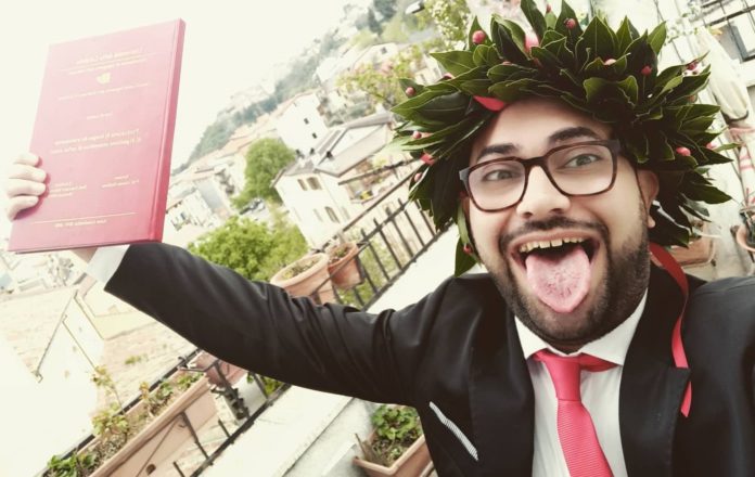 Laurea Francesco SPina nella foto è seduto alla scrivania, con le spalle rivolte al computer. Tiene in mano la cartellina della laurea. Indossa camicia bianca, cravatta rossa e giacca nera. Sulla testa indossa una corona d'alloro da laureato, indossa occhiali da vista, ha la brba e sorride felice facendo la linguaccia