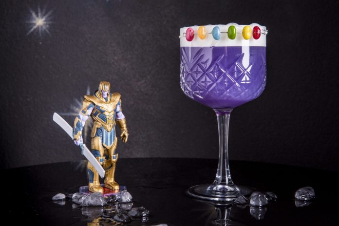 Thanos Infinity Gauntlet - nella foto il drink in una coppa viola con delle caramelline rotonde colorate intorno al bordo del bicchiere, tenute su da un filo su una striscia bianca, e affianco il modellino del personaggio Thanos con l'armatura d'oro, i guanti magici e in mano una lancia composta da due pale bianche