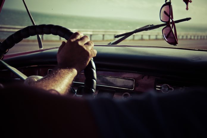 Primo piano del lunotto di un'auto con un volante, una mano di uomo appoggiata sopra, i tergicristalli e appesi allo specchietto, un paio di occhiali da sole