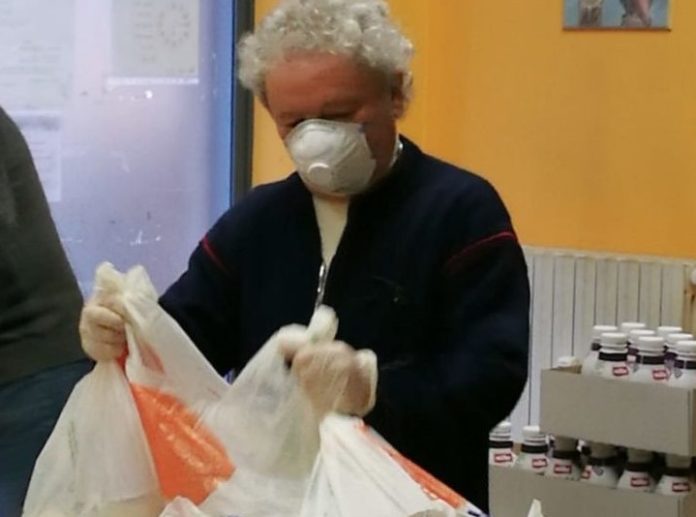 Pasqua in povertà - Don Adriano, con abiti neri, capelli bianchi e mascherina bianca, prepara delle borse di derrate alimentari per i poveri
