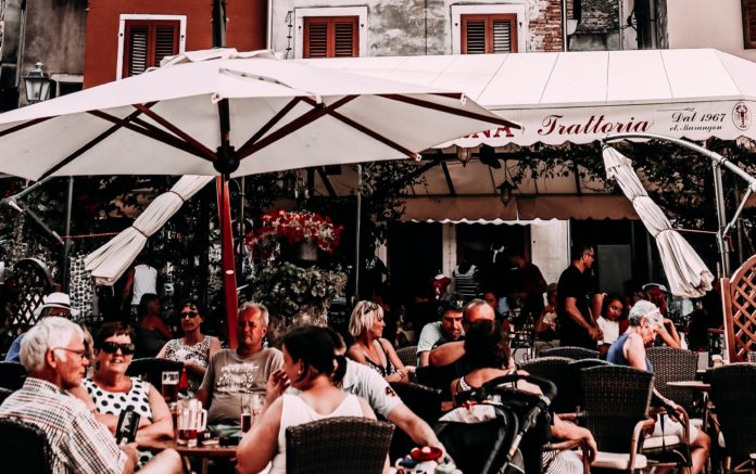 Ristorazione - una piazza con dei tavolini di ristoranti con tantagente seduta ai tavoli coperti da grandi ombrelloni bianchi