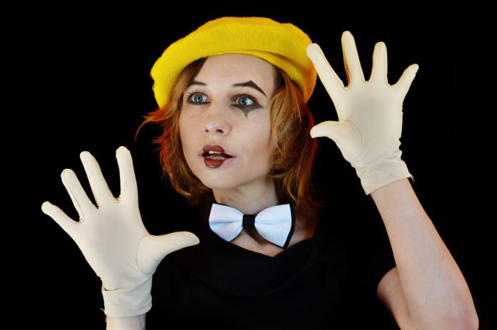 Roma Fringe Festival 2021 - una ragazza con la faccia truccata di bianco,, cn capelli rossi acaschetto, indossa un cappello giallo e dei guanti bianchi. tiene le mani davanti al viso ma senza coprirlo, con un'espressione di stupore. Al collo ha un farfallino bianco su maglietta nera