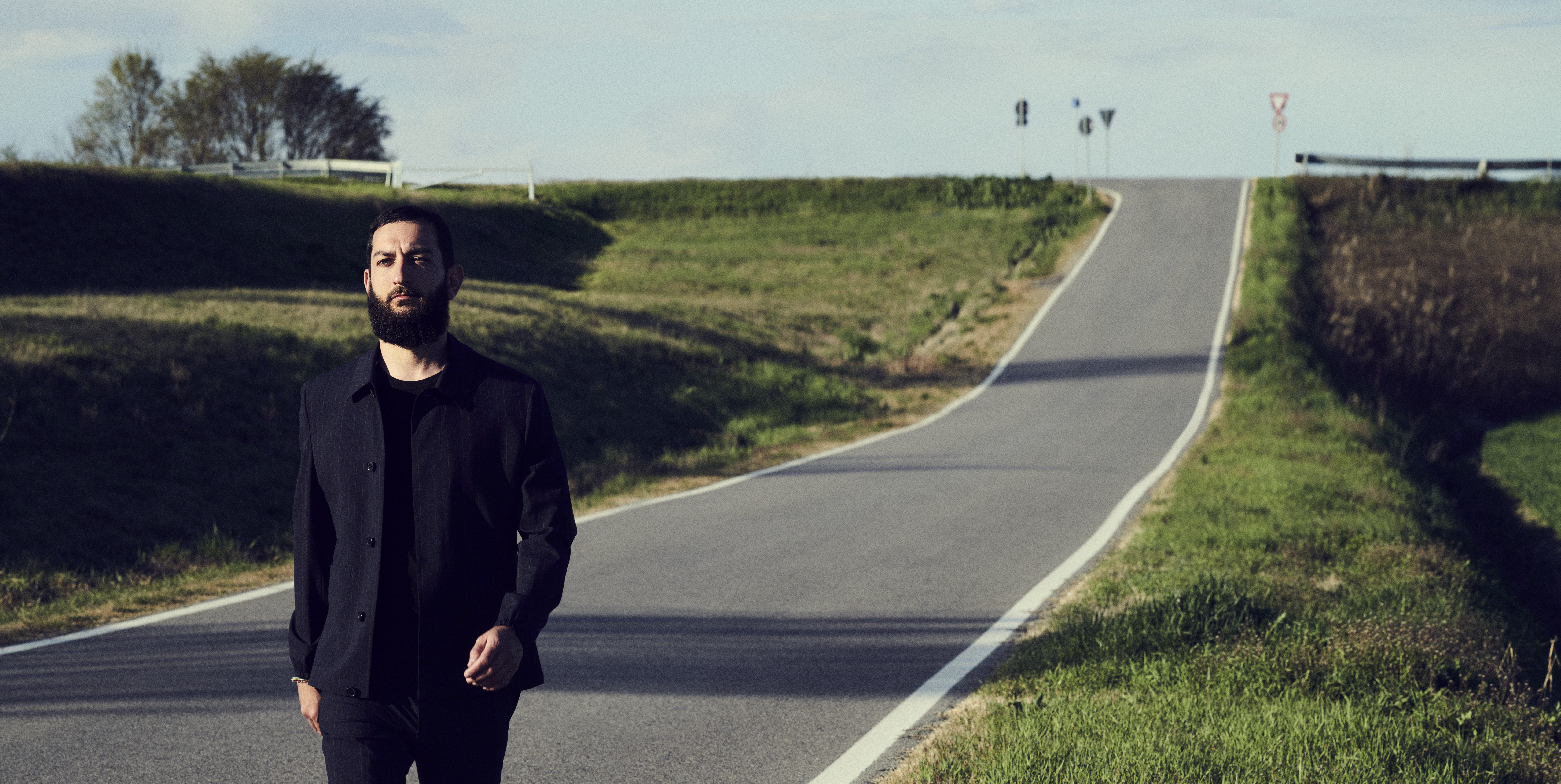 Vasco Brondi in foto con una strada collinare ferrarese solo sfondo, vestito di nero al centro, il cielo sopra azzurro