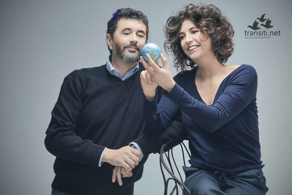 TRansiti - Anna-Pisterzi-e-Mauro-Sclavo seduti vicini vestiti entrambi con un maglione scuro, guardano sorridendo un piccolo mondo tenuto in mano da Anna
