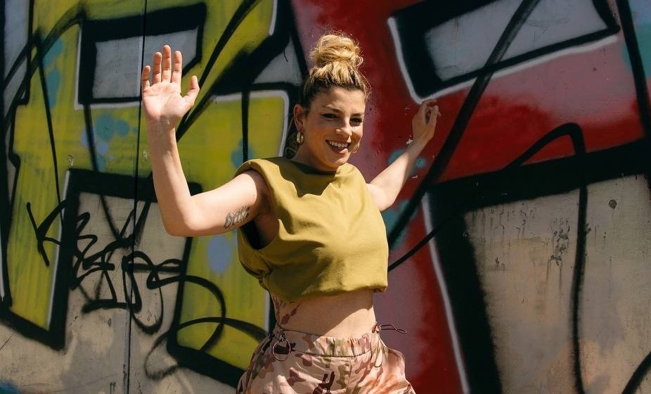 Emma con t shirt tagliata all'ombelico, alza le braccia sorridente davanti a un murales