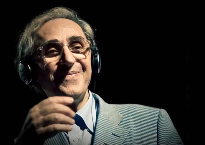 Franco Battiato con giacca grigia, camicia grigia, con cuffie da musica in testa sorride