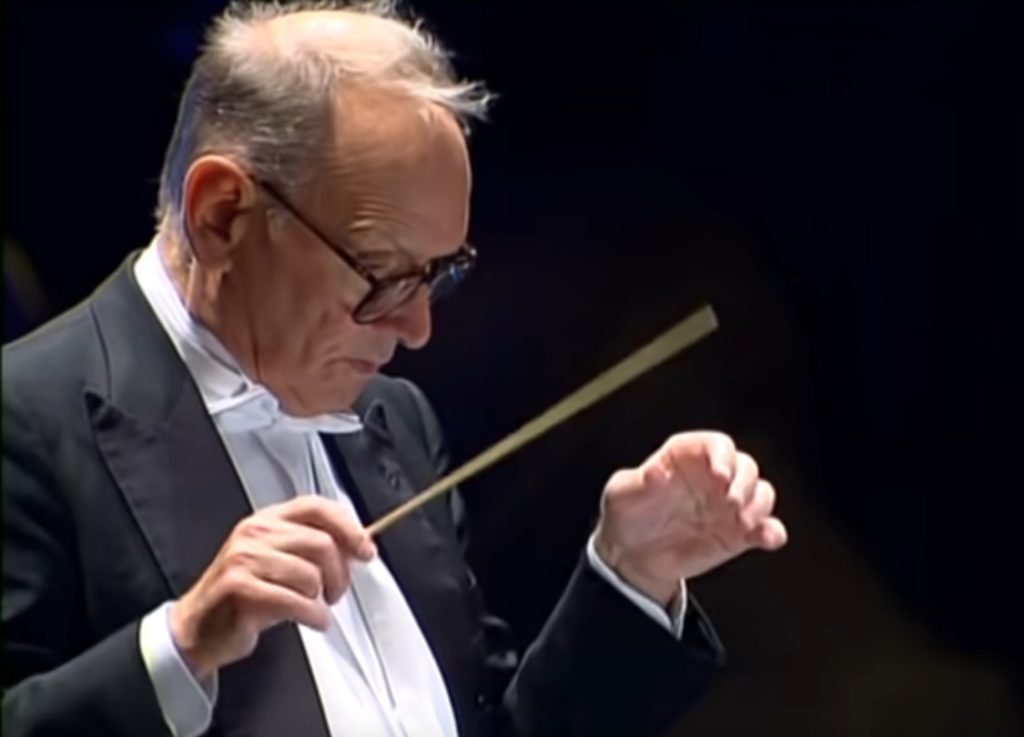 Ennio Morricone, anziano, di profilo, con occhiali da vista con montatura scura, indossa camicia bianca e giacca nera, sta dirigendo un'orchestra con la bacchetta nella mano destra