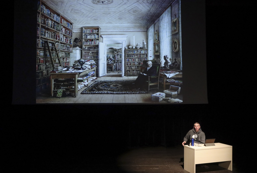 QuinteScienza  - Un frame dello spettacolo con un uomo seduto ad un tavolo sulla destra e nel buio solo la proiezione su uno schermo gigante di una stanza-libreria con tanti libri sugli scaffali, un tappeto al centro del pavimento di legno ed un uomo anziano con la barba seduto su una poltrona, ai suoi piedi delle casse, una aperta l'altra chiusa