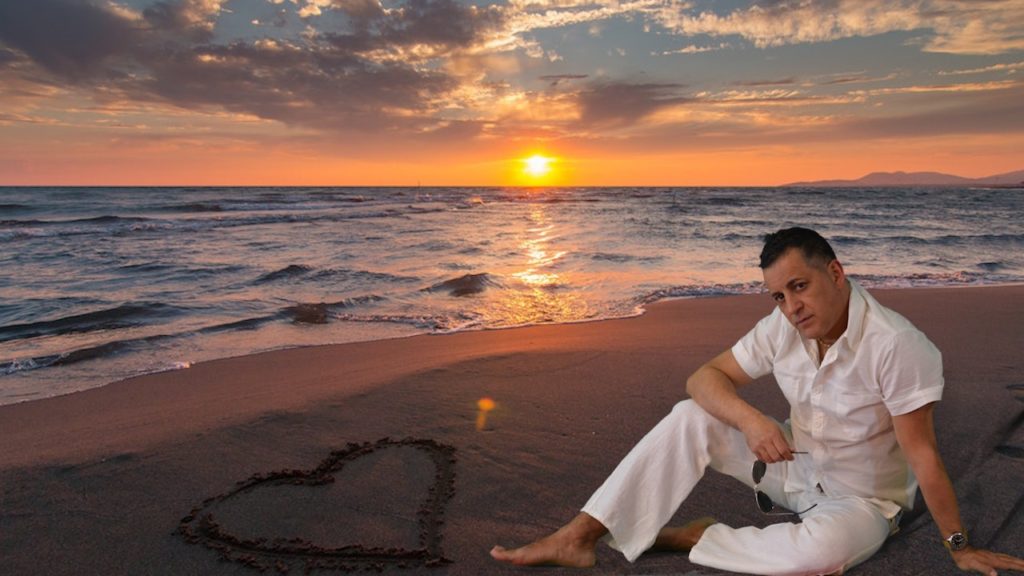 Massimiliano Balocchi vestito di bianco, seduto su una spiaggia in riva al mare con un bellissimo tramonto, sulla sabbia c'è un cuore disegnato
