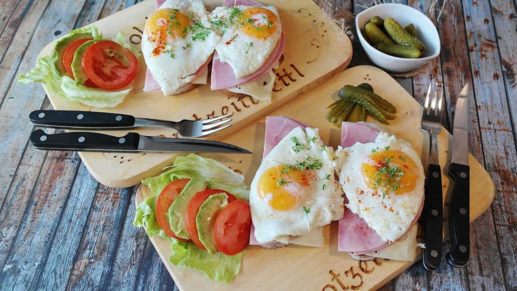 social e food porn - due taglieri di legno rettangolari con delle uova, del prosciutto e dei pomodorini e dell'insalata. Su uno di quest sono appoggiate delle posate