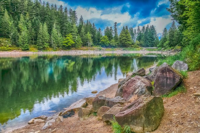 Lago delle fate, gnomi. Lago con acqua cristallina, di colore verde azzurro riflette l'immagine del cielo caratterizzato dalla presenza di nuvole. Intorno vasta vegetazione di colore verde acceso.