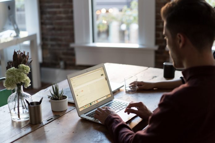 Lavoro agile - un ragazzo dseduto al tavolo della sua cucina sta lavorando al computer con una tazza di caffè sulla destra, uuna piantina davanti a lui, dietro ilmonitor del pc e sullo sfondo una finestra