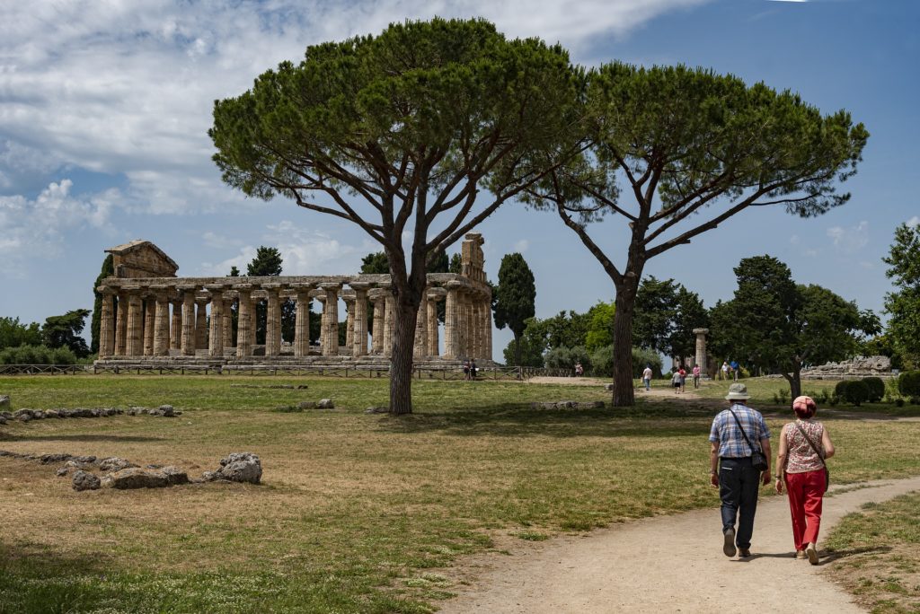 Paestum il tempio visto da lontano , con un prato, le rovine del tempio fatto di colonne. Sulla destra, una coppia di turisti passeggia sulla strada sterrata