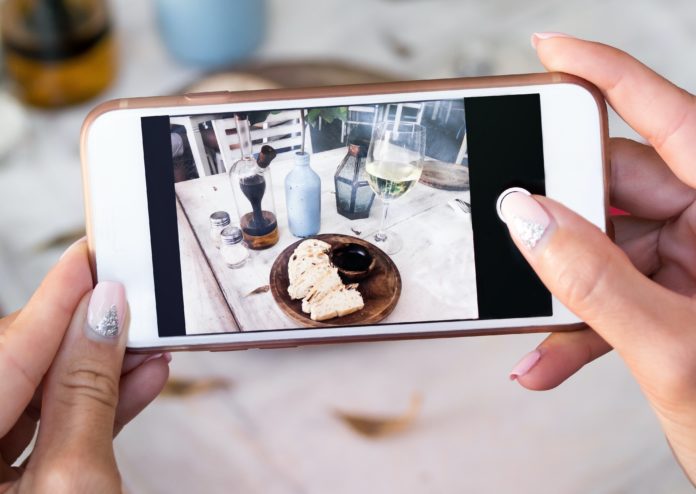 food porn due mani tengono un telefonino, intente a fotografa del cibo. sulla tavola che stanno fotografando ci sono bicchieri, bottiglie di vino e di acqua, un piatto con del cibo