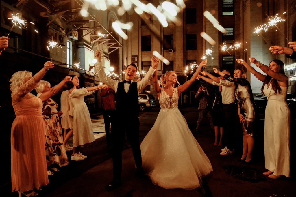 Due sposi fanno la passerella tra gli invitati. La sposa ha un bellissimo abito bianco lungo e llargo al fondo, lo sposo, ha vestito scuro, entrambi hanno le braccia alzate.