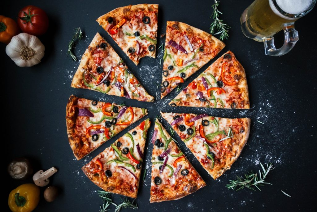 Una pizza capricciosa, con peperoni a listelle, olive, mozzarella cipolle, divisa a spicchi, vista dall'alto, appoggiata su un tavolo nero, con intorno dei rametti di rosmarino, dei peperoni, dell'aglio