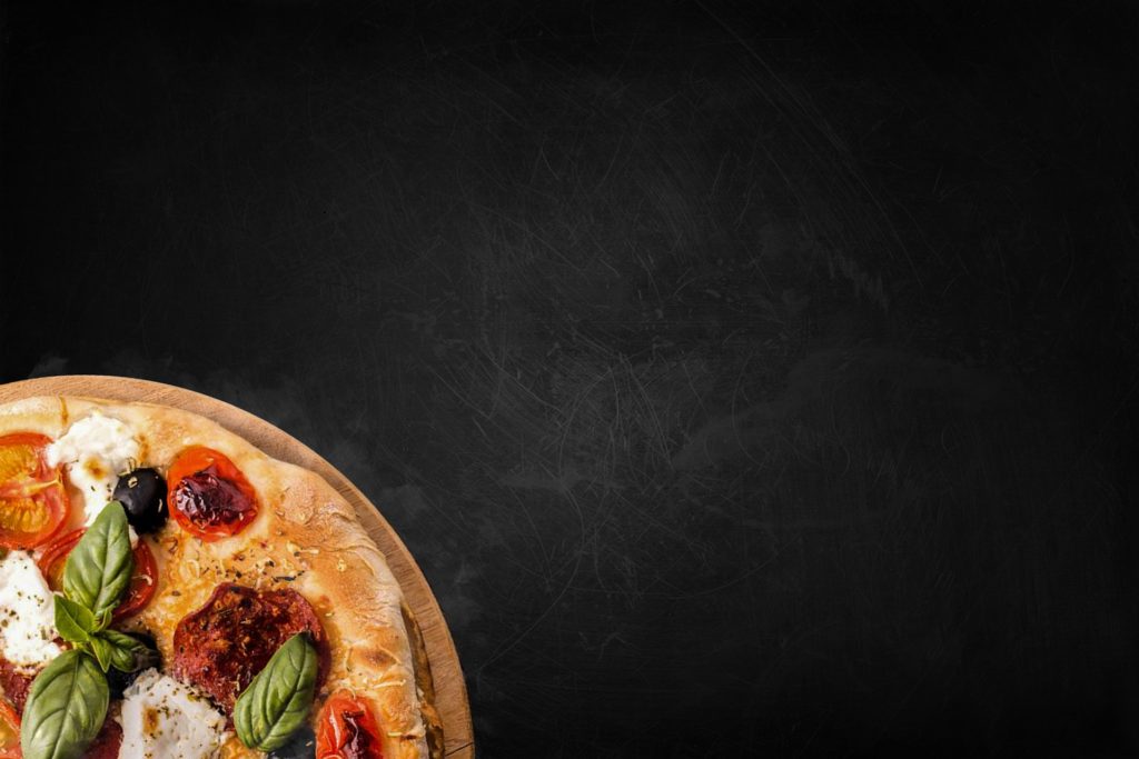 uno sfondo nero con un quarto di pizza in basso a destra. Sulla pizza c'è del pomodoro a fette, delle foglie di basilico e della mozzarella fusa