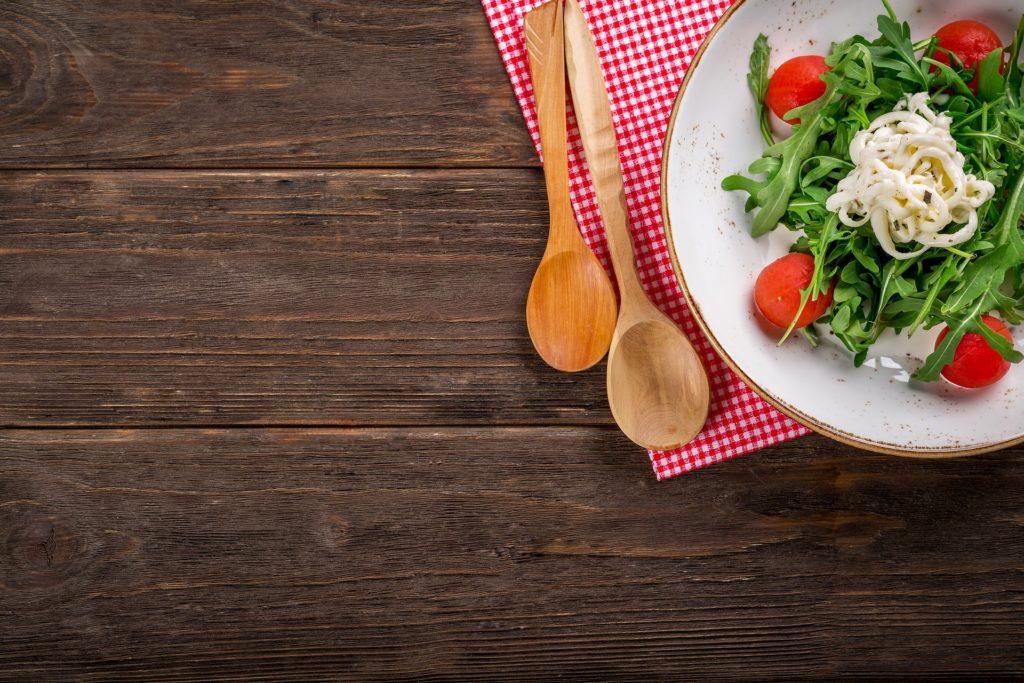 cibo su un tavolo di legno, sulla sinistra si vede una parte di una tovaglietta rossa a quadretti con sopra un piatto bianco cin della verdura e dei pomodori e vicino al pitto due cucchiai di legno
