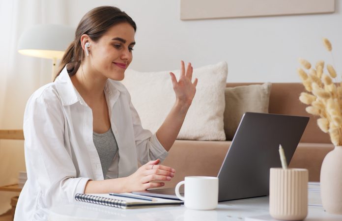 smart working - una donna ben curata, con capelli biondi legati, camicia bianca aperta su una t shirt verde, è davanti ad un pc portatile e saluta con la mano sinistra. Sul tavolo bianco, oltre al pc, ci sono una tazza bianca, un vaso di fiori e un porta penne cilindrico. Sullo sfondo si vede un divano e dei cuscinoni