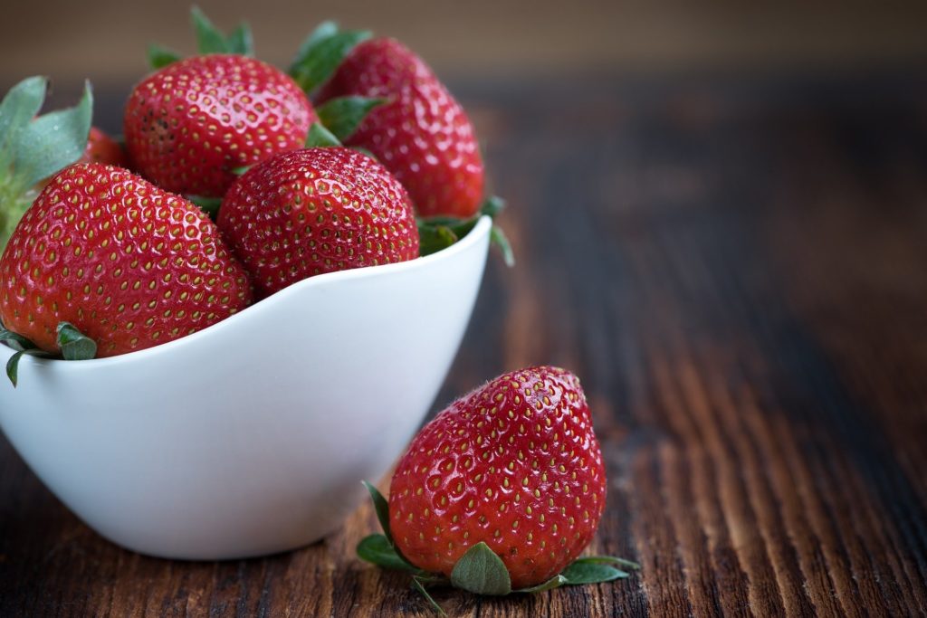 food porn :sulla sinistra, una ciotola bianca contenente delle fragole, fuori dalla ciotola, ma vicina una fragola rossa grossa, su un tavolo di legnole