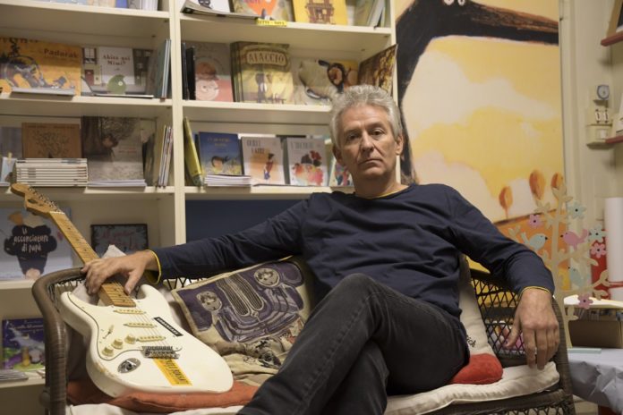 paolo bonfanti, il bluesman di Genova in foto seduto sul divano, dietro lo scaffale con i libri, accanto a lui la chitarra