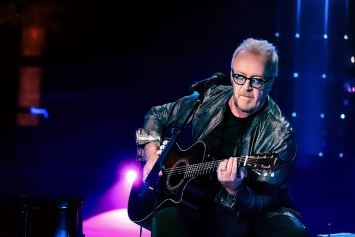 Umberto Tozzi in un concerto, veste giacca dipelle nera, è seduto su uno sgabello e suona la chitarra. Intorno a lui le luci sono viola