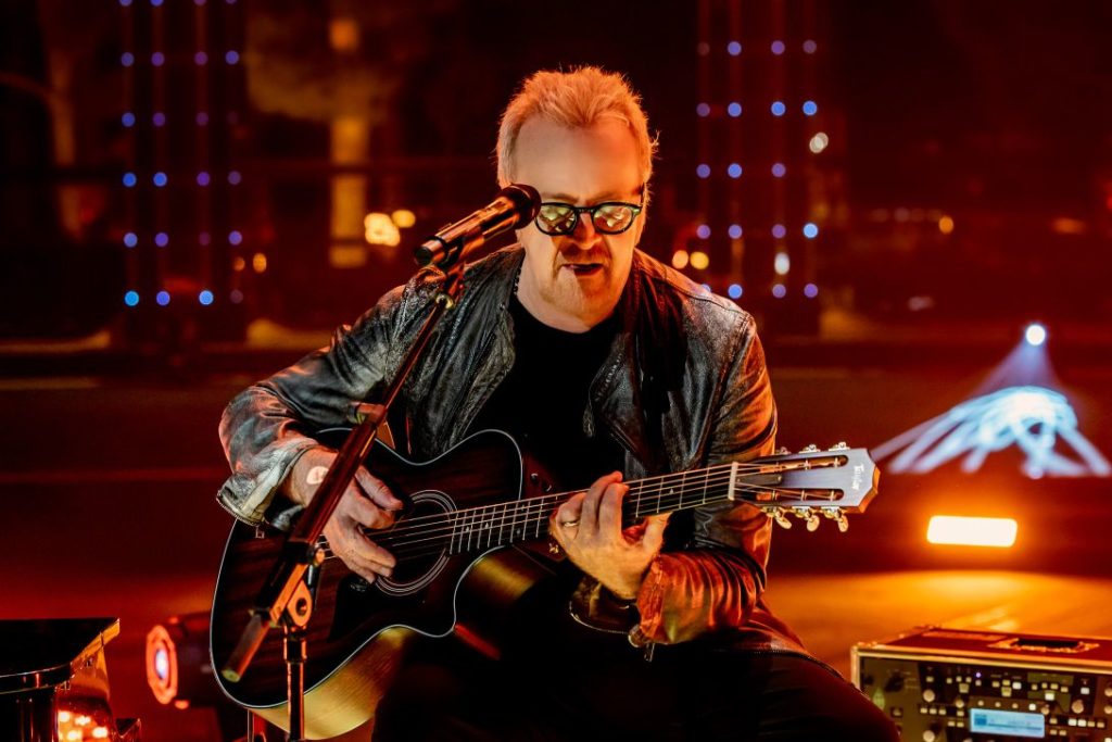 Umberto Tozzi in un concerto, veste giacca dipelle nera, è seduto su uno sgabello e suona la chitarra. Intorno a lui le luci sono arancioni