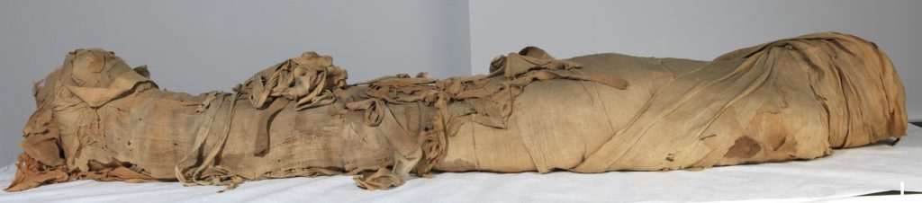 una mummmia distesa su un lettino, fatta con diversi tessuti e bende di colore beige