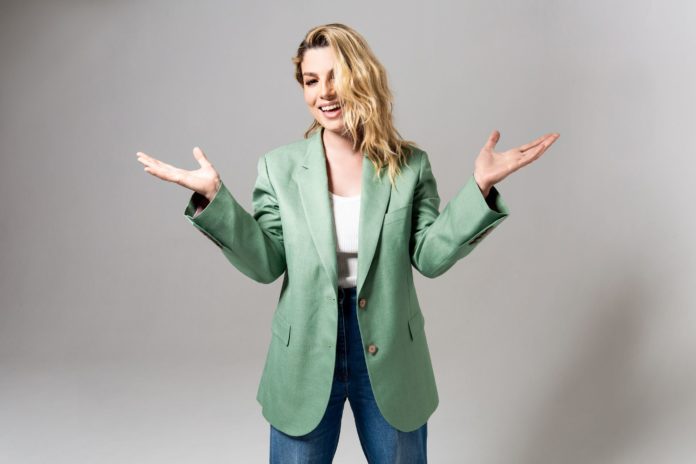 Emma live all'Arena di Verona veste un completo di giacca verde e jeans, con una t shirt bianca e tiene le braccia aperte. Ha lunghi capelli biondi e sorride