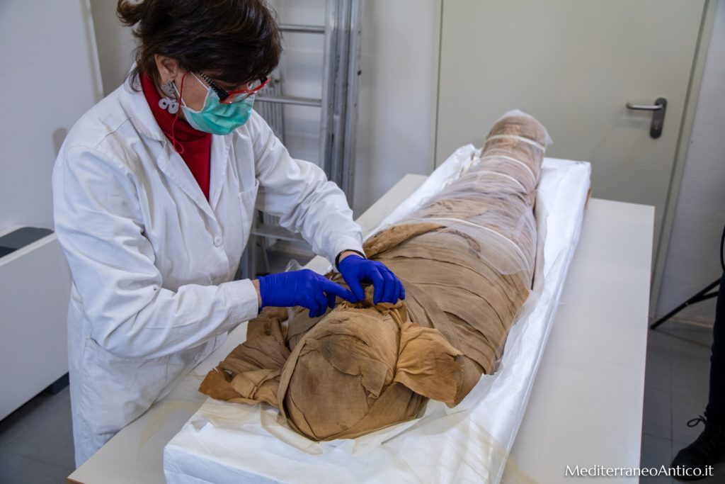 una restauratrice sta operando su una mimmia egiia di colore beige, distesa su un lettino bianco all'interno di un laborartorio