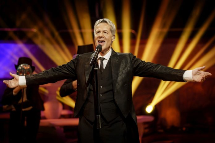 Claudio Baglioni vestito con giacca e cravatta, gillet, ha le braccia aperte e canta davanti a un microfono