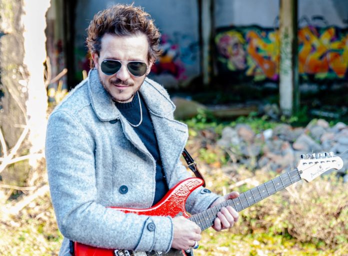 indelebile - demis facchinetti, giaccone di lana invernale di colore chiaro, occhiali da sole, con una chitarra elettrica rossa a tracolla
