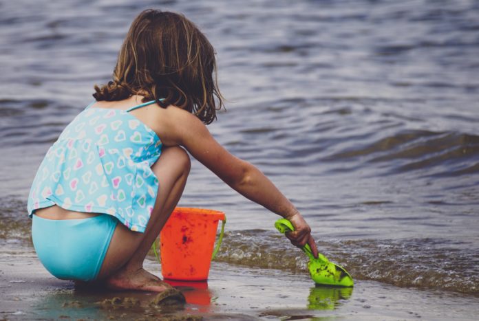 granchio nel secchiello - una bambino accovacciata sulle ginocchia, in riva al mare raccoglie sabbia con un apaletta verde per metterla in un secchiello rosso. La bambina indossa una canottiera a fiori azzurra e dei pantaloncini azzurri ed ha lunghi capelli neri