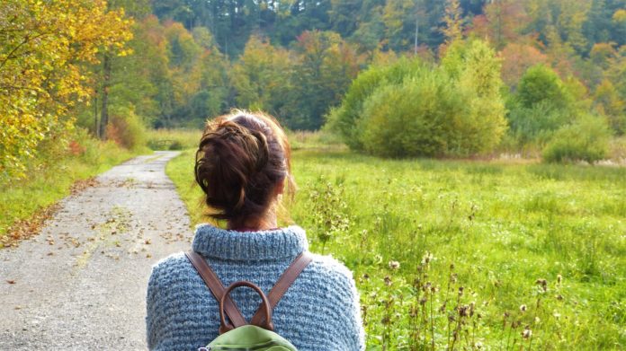 green pass: una donna di spalle con capelli raccolti sulla nuca, indossa un cardigan azzurro e ha uno zainetto sulle spalle. la donna cammina in mezzo ad una campagna ricca di alberi e prati verdi