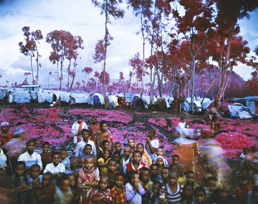 Richard Mosse in questa foto ha immortalato un gruppo di bambini congolesi in un campo profughi. Sullo sfondo diverse abitazioni fatte di tende. La foto presenta ogni cosa con tonalità di rosso e rosa