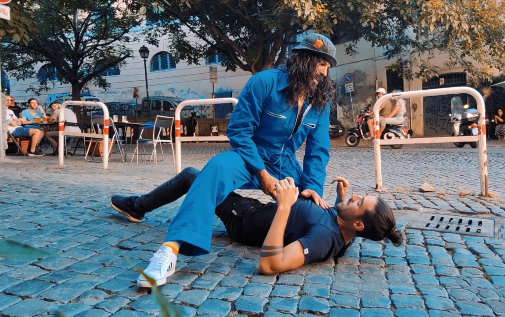 Tomas Milian - nella foto Rocky atterra con un amossa da wrestling un uomo con capelli raccolti, vestito con maglia blu e pantaloni neri. Rocky indossa tuta blu da meccanico e cappello, con barba e capelli neri