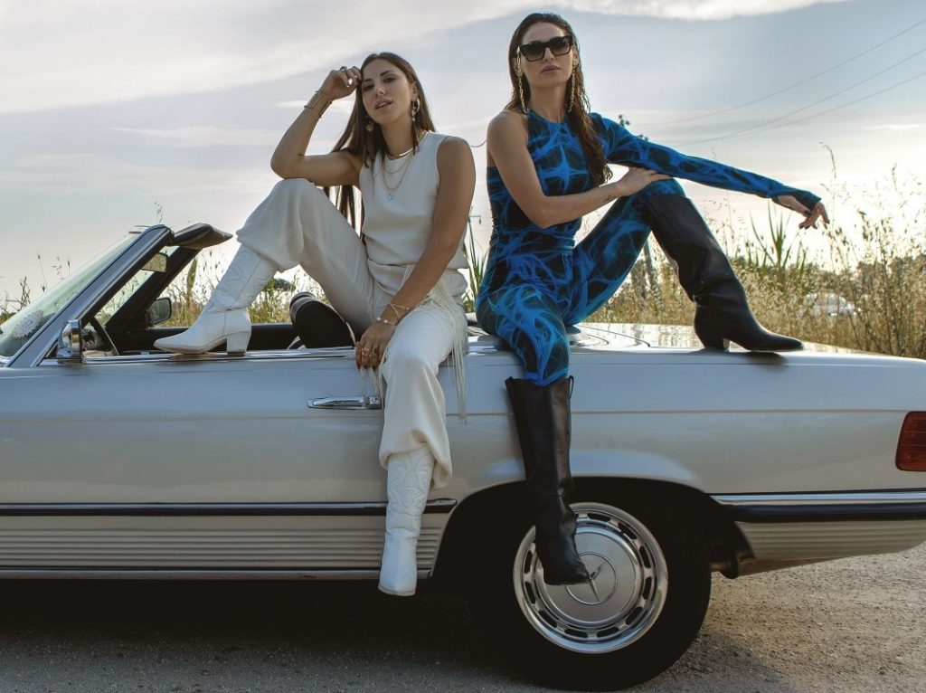 Anna Tatangelo con Beba. Le due ragazze sono sedute sul bordo della carrozzeria di una macchiana con la capotte abbassata. Beba, a sinistra, indossa un completo canottiera e pantalone bianco e ha la mano appoggiata sulla fronte, Anna, a destra, indossa una tuta blu attillata, stivaloni neri e ha una mano appoggiata sul ginocchio