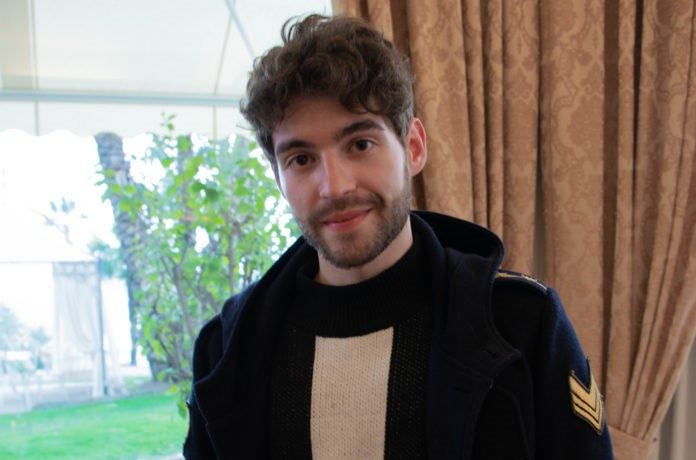 Matteo Faustini inprimo piano veste un giaccone nero, un maglione beige, ha capelli e barba neri e soprride. E' in una stanza con delle tende pesanti arancioni e dalla finestra si vedono degli alberi