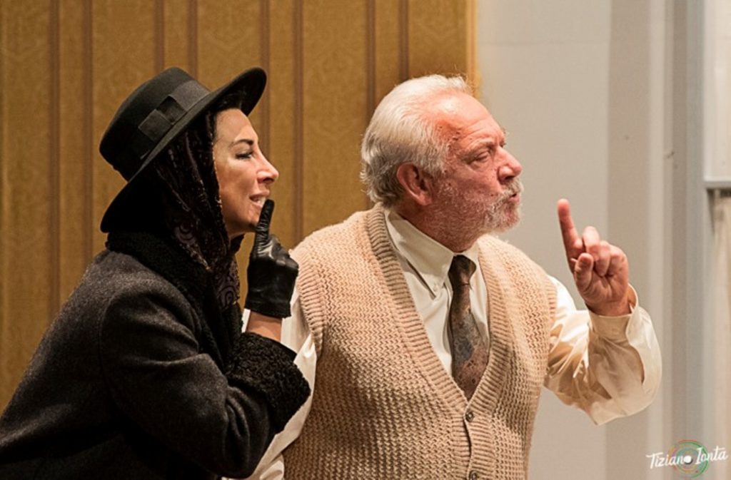 Piombino - Nella foto una donna, a sinistra, con cappotto e cappello nero, solleva un dito verso la bocca in segno di silenzio. La mano è coperta da un guanto nero. Sulla sua destra un uomo anziano con capelli bianch, indossa una camicia bianca una cravatta marrone e un gillet beige e ache lui con il dito fa il segno di silenzio