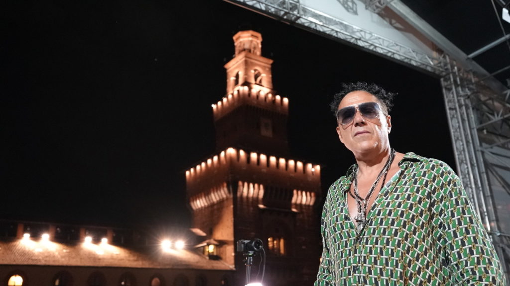 Ferragosto a MIlano - nella foto il dj Joe T Vannelli con camicia verde a quadri, occhiali scuri, e sullo sfondo il castello sforzesco di Mialno di notte, illuminato da luci suggestive
